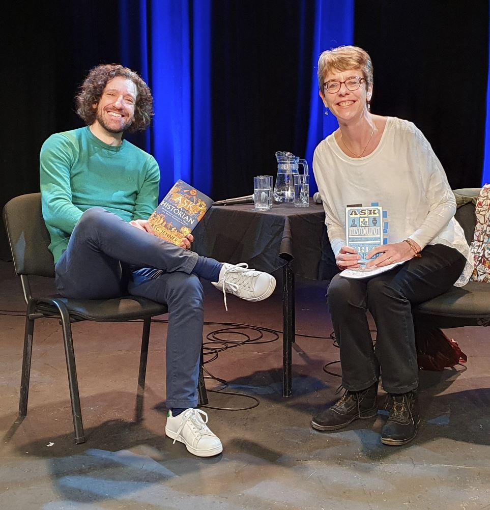 Greg Jenner and Ally Sherrick sitting on the stage with copies of Greg's new book